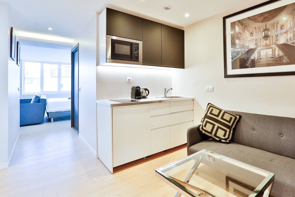 A modern kitchen with a microwave, sink, and coffee maker. There is a glass table and a sofa with a patterned pillow. A framed picture hangs on the wall.
