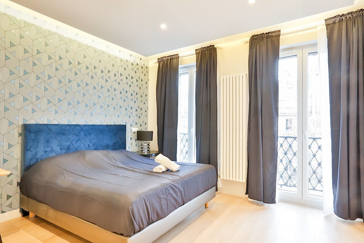A modern bedroom with a large bed, a bedside table with a lamp, and a view of the outside through large windows with curtains.