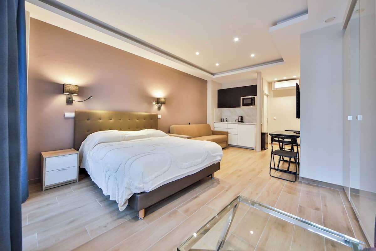 A modern bedroom with a large bed, bedside tables, a sofa, and a kitchen area in the background.