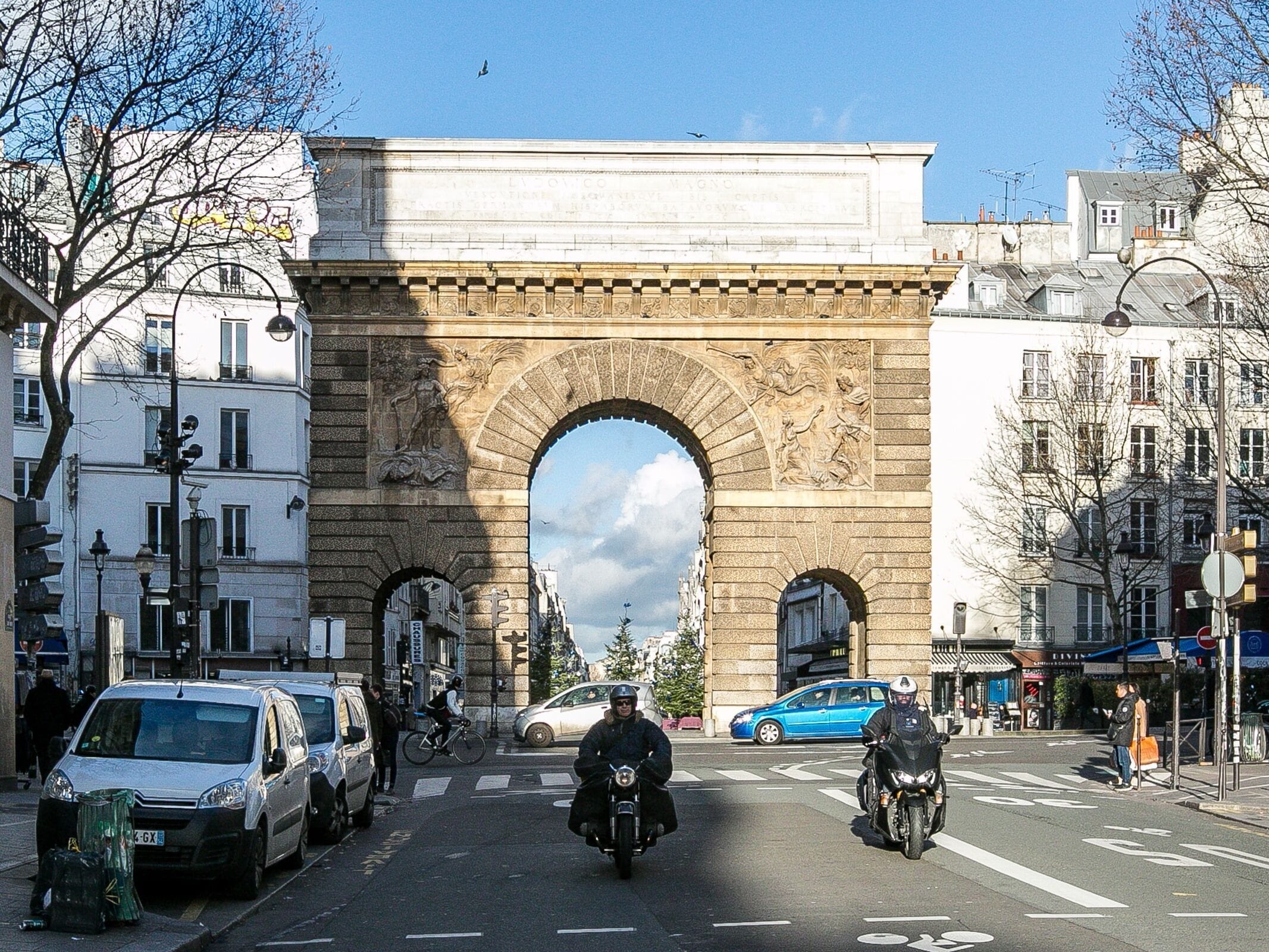 A street scene with a large stone archway, vehicles, and buildings.