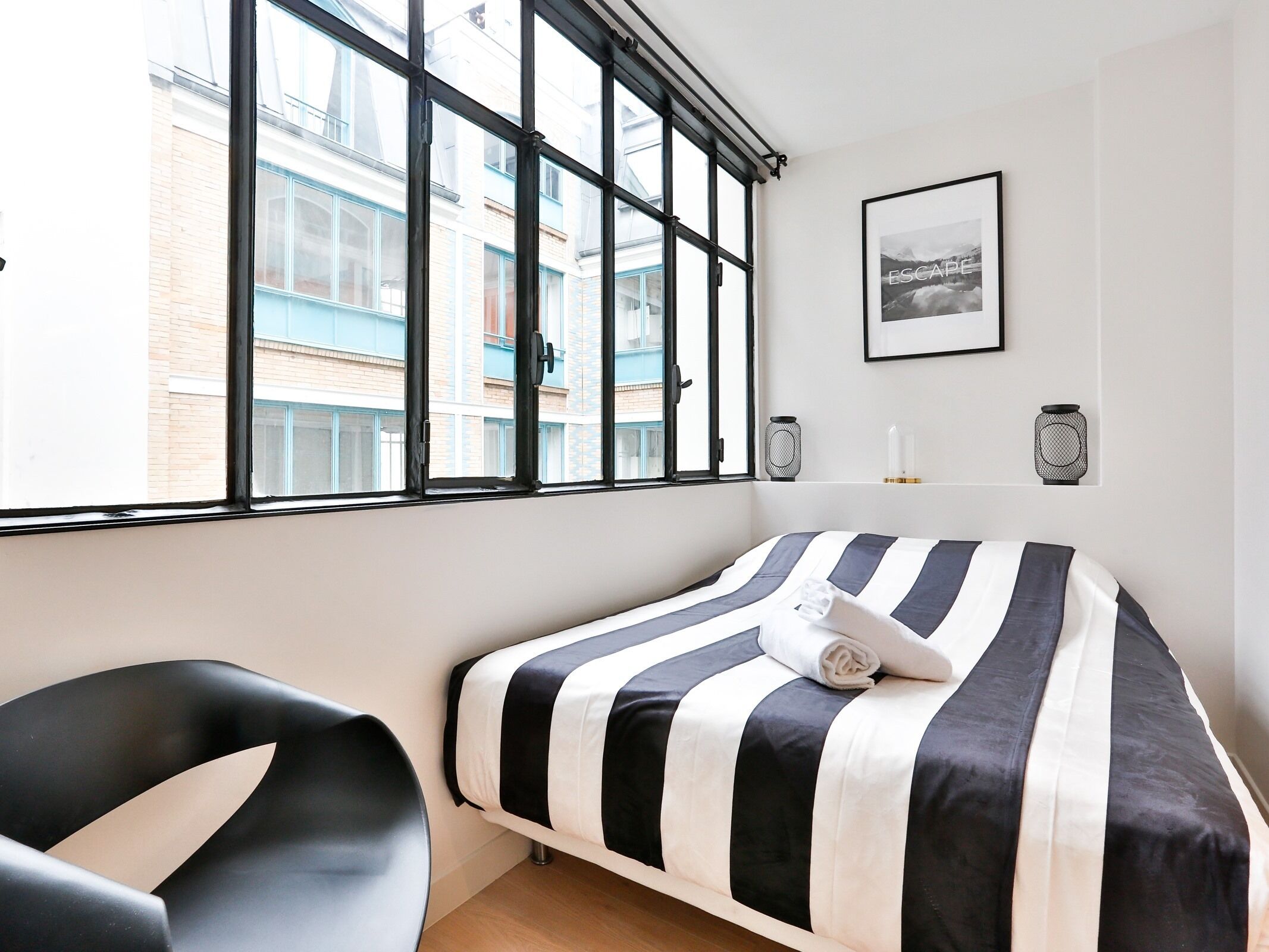 A neatly arranged bedroom with a bed, a chair, and a large window offering a view of buildings outside.