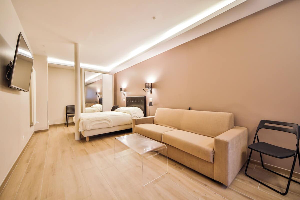 A modern hotel room with a bed, a sofa, a small table, and a TV mounted on the wall.