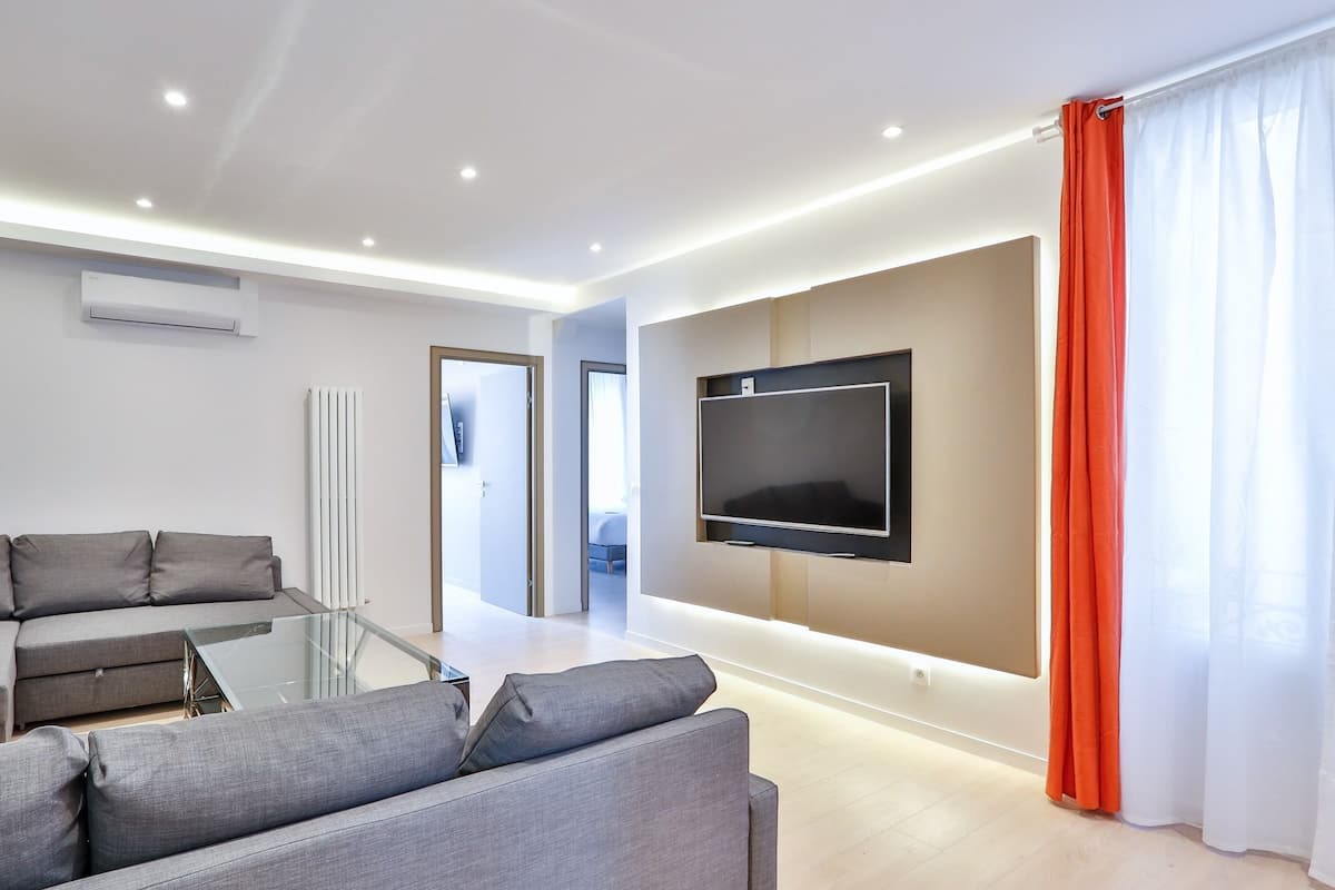A modern living room with a grey sofa, a flat-screen TV mounted on the wall, and a glass coffee table.