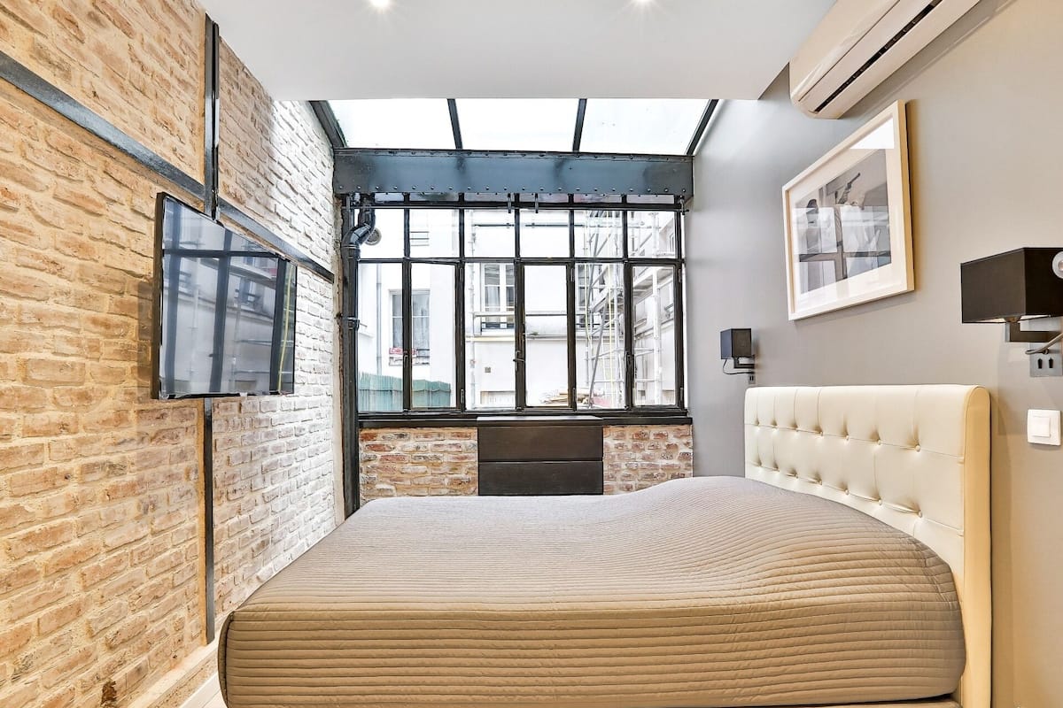 A bedroom with a large bed, a TV mounted on the wall, a window with a view of the outside, and a brick wall.