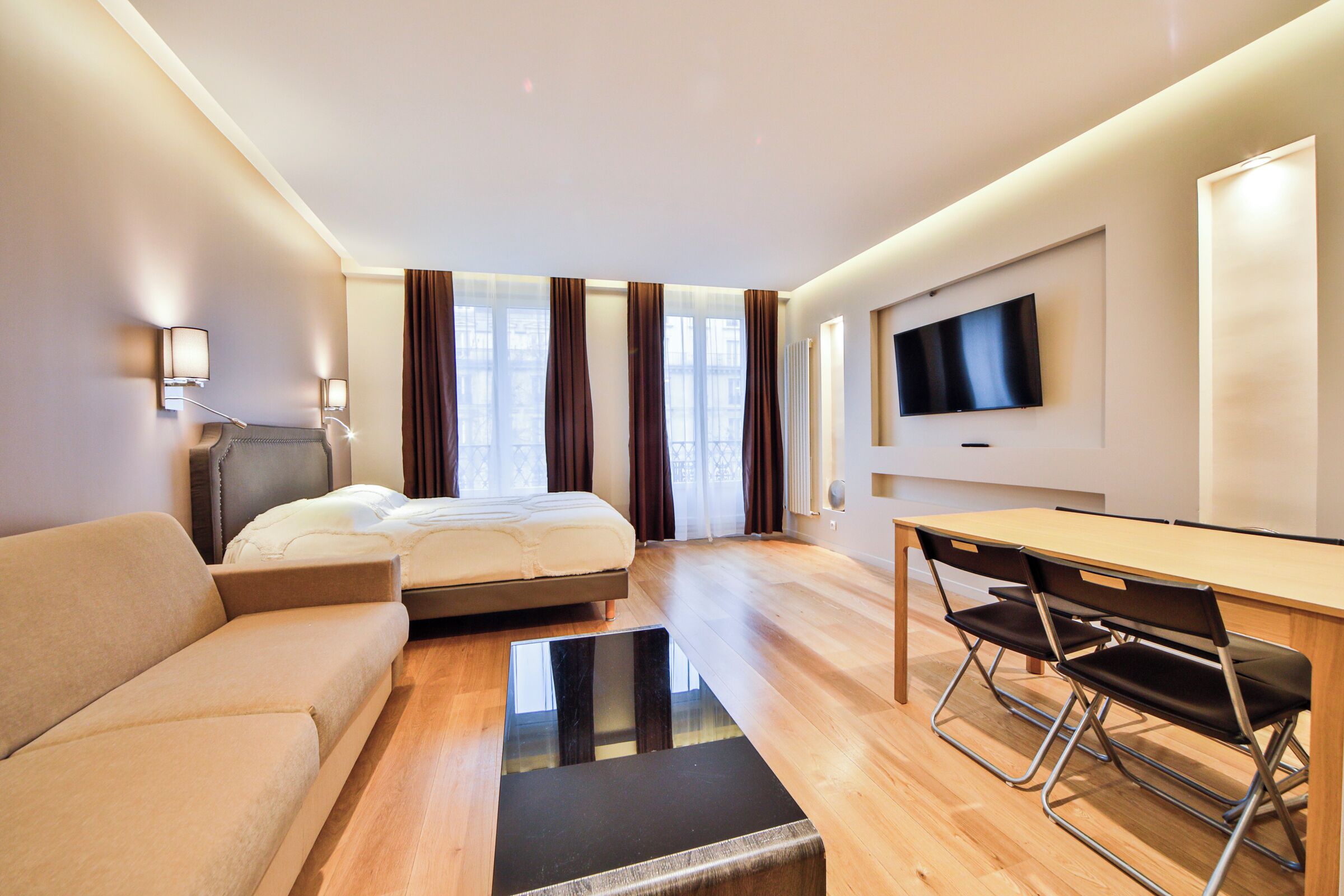 A modern hotel room with a bed, sofa, desk, chairs, and a TV on the wall.