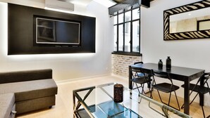 Een moderne woonkamer met een flatscreen televisie aan de muur, een glazen salontafel, een bank en een eethoek met een tafel en stoelen.