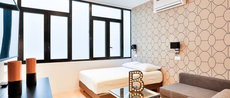 A modern hotel room with a bed, a glass coffee table, a sofa, and a large window with blinds.