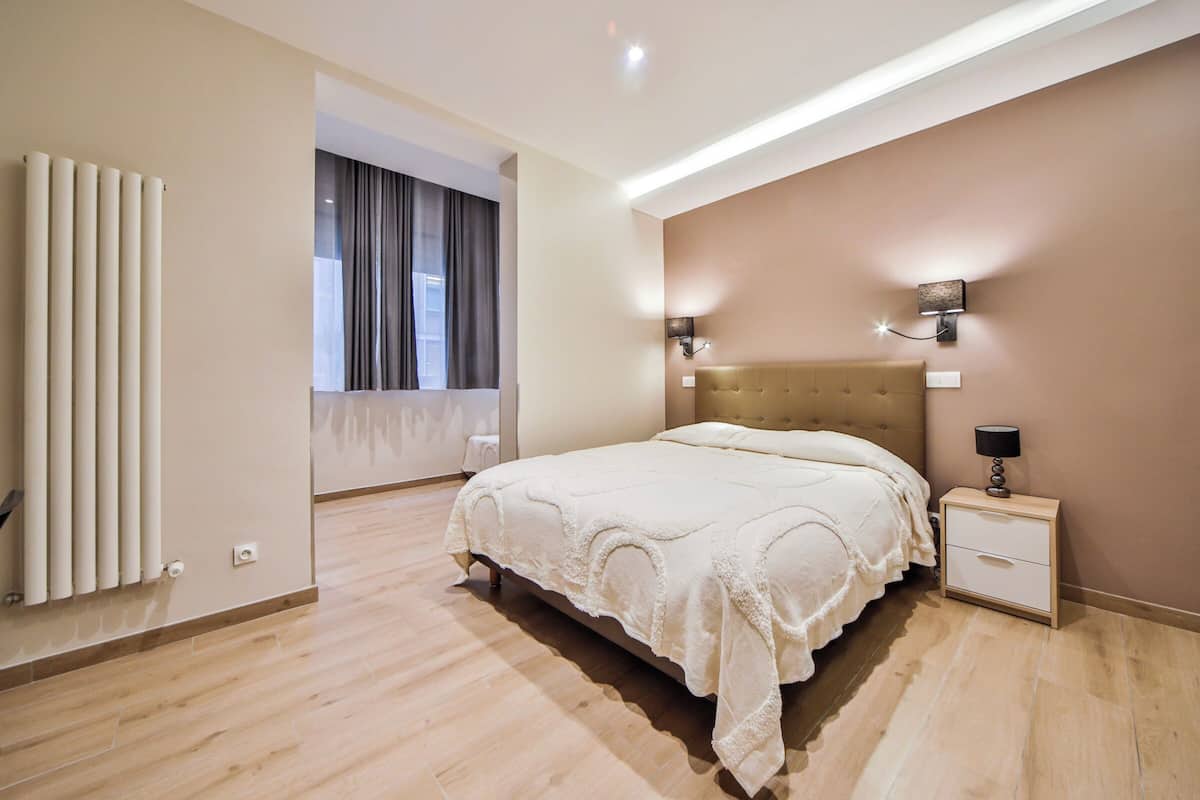 A modern bedroom with a large bed, a nightstand, a wall-mounted lamp, and a radiator.
