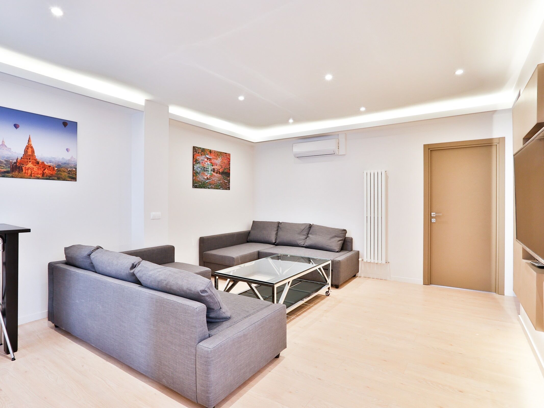 A modern living room with two grey sofas, a glass coffee table, and a wall-mounted air conditioner.