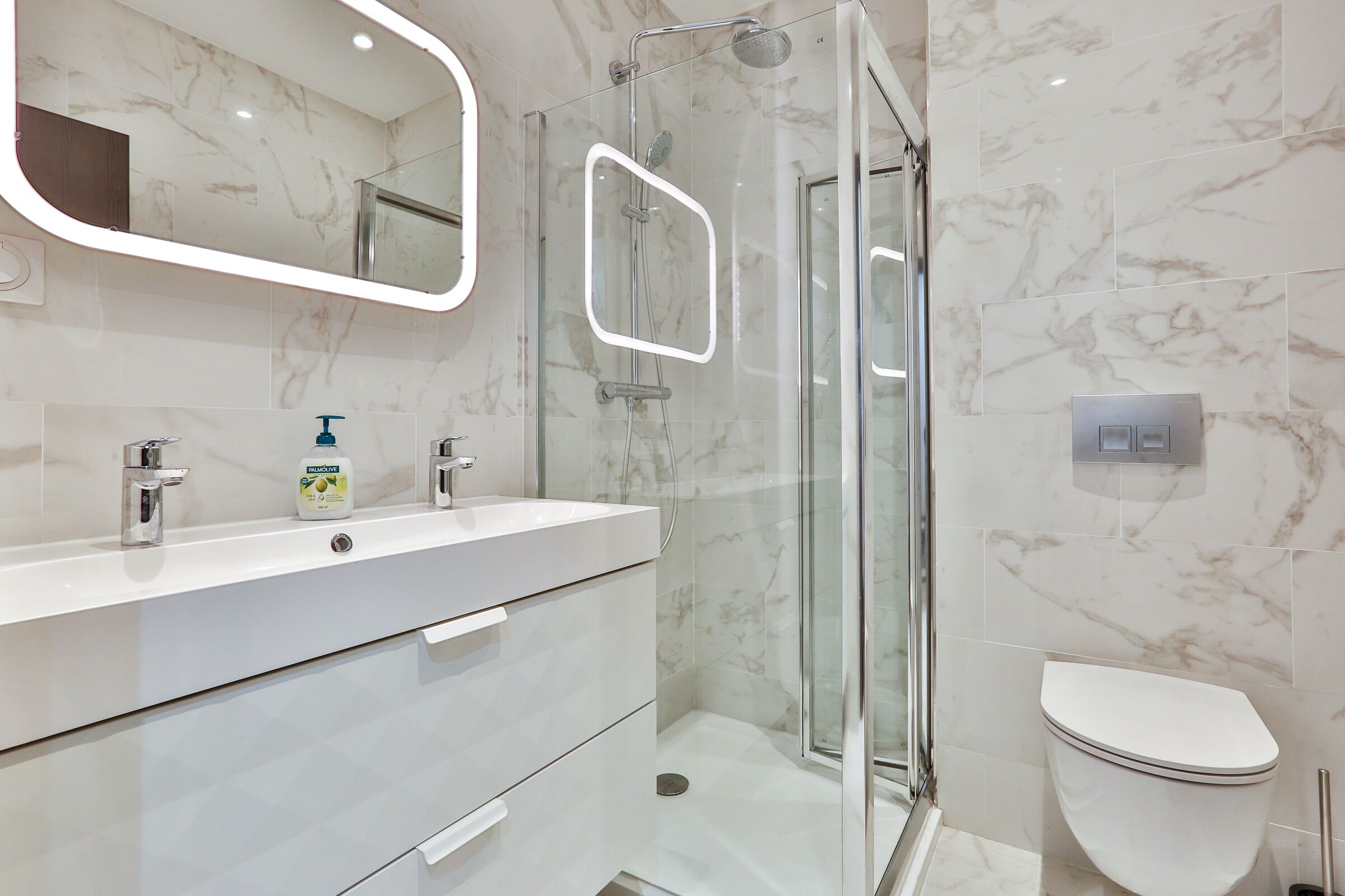 Un baño moderno con ducha de vidrio, lavamanos blanco con dispensador de jabón y un inodoro.