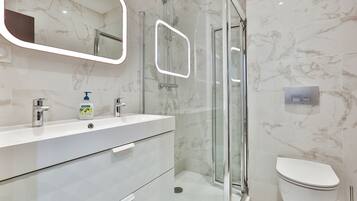 Un baño moderno con ducha de vidrio, lavamanos blanco con dispensador de jabón y un inodoro.