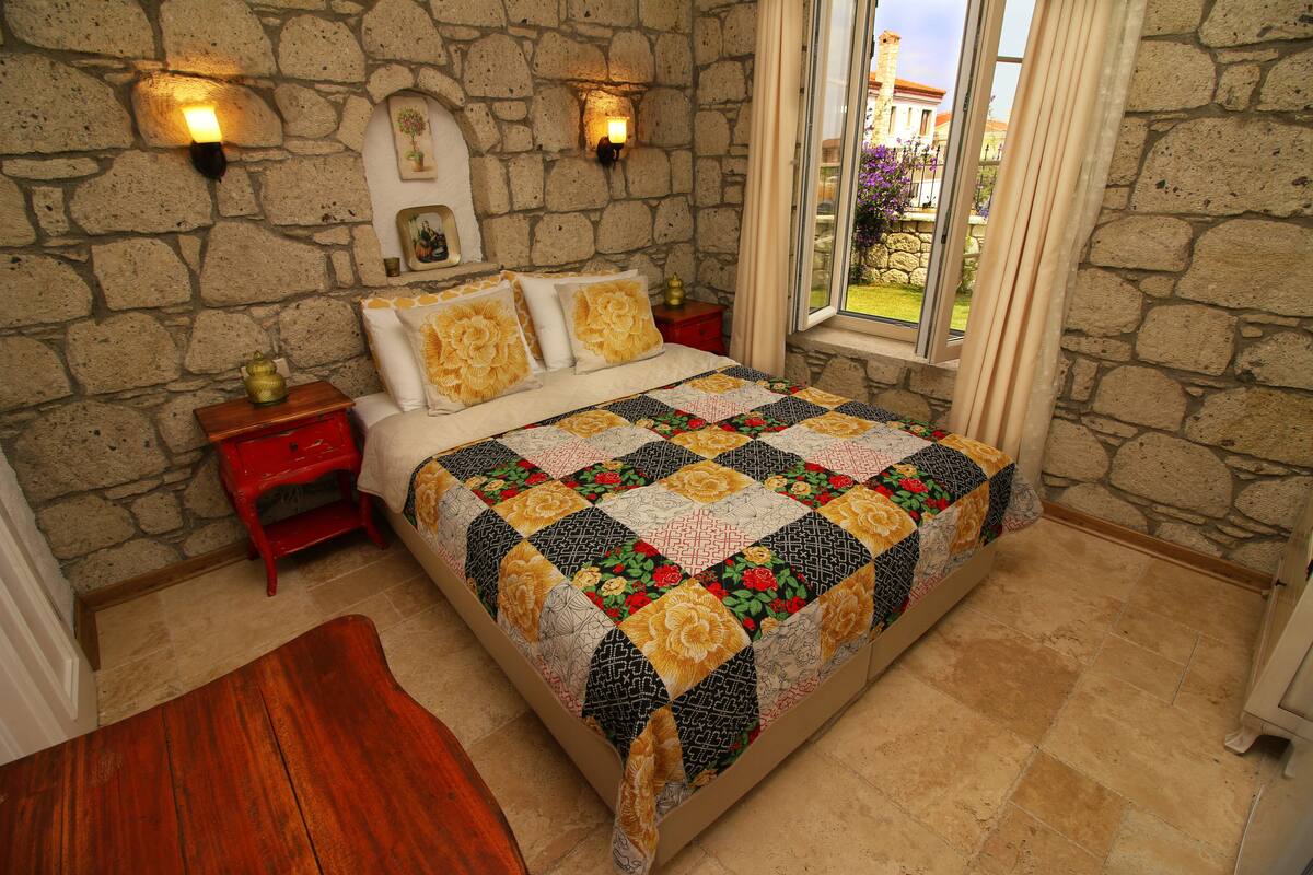 A bedroom with a stone wall, a wooden floor, a bed with a quilt, two wall-mounted lights, and a window with curtains.