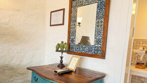 A blue wooden dresser with drawers, a framed mirror with a decorative frame, a wall-mounted light fixture, and a small shelf with decorative items.