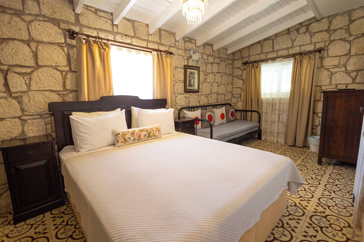 A bedroom with a large bed, a wooden nightstand, a dresser, and a stone wall.