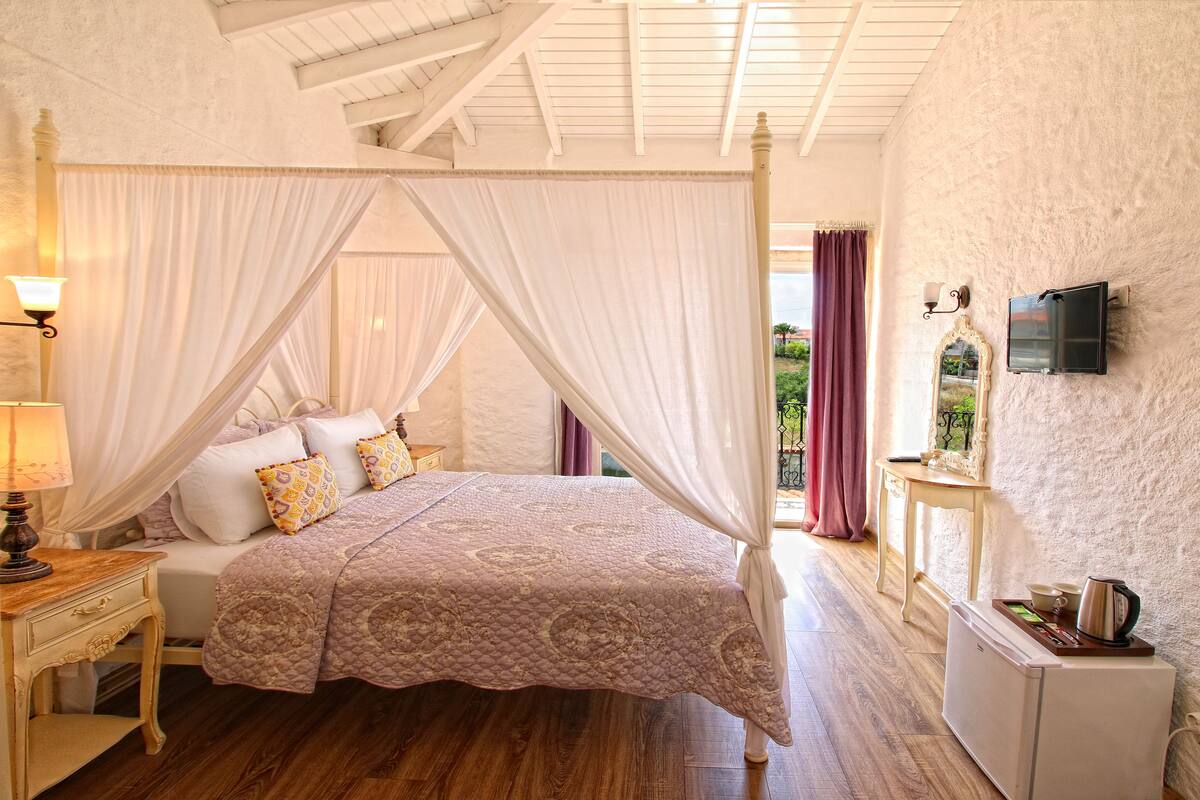 A bedroom with a four-poster bed, a bedside table with a lamp, a small white cabinet, and a television mounted on the wall.