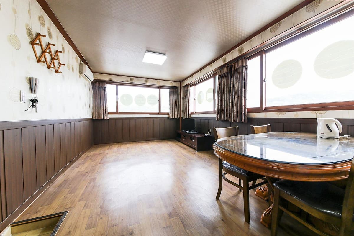 A room with a wooden floor, a round glass table with chairs, a television, and a kettle on the table.