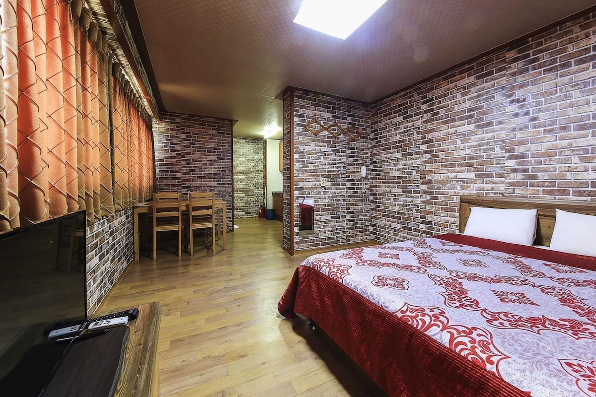 A room with a brick wall, a bed with a red and white patterned bedspread, a wooden desk with a chair, and a skylight.