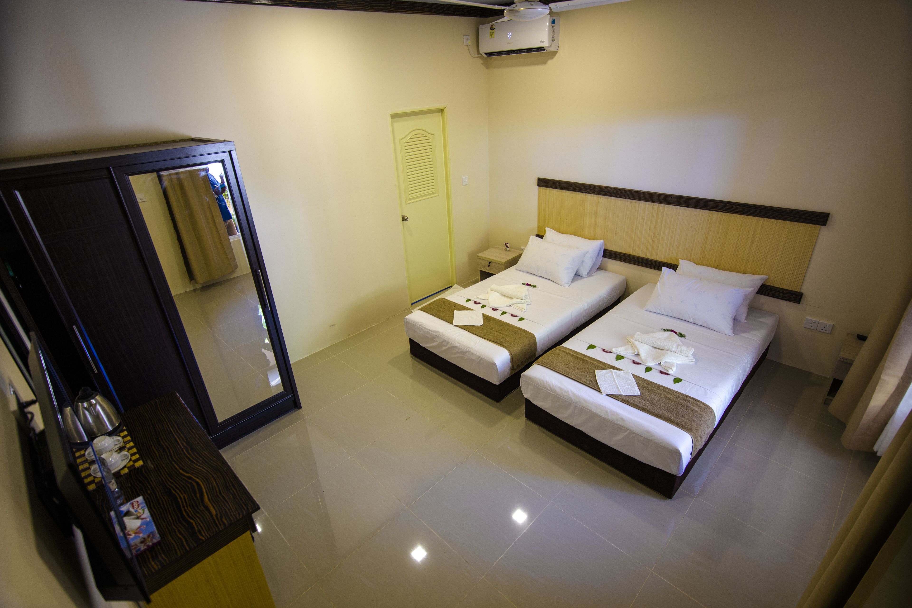 A hotel room with two beds, a wardrobe, a bedside table with a kettle, and a mirror.
