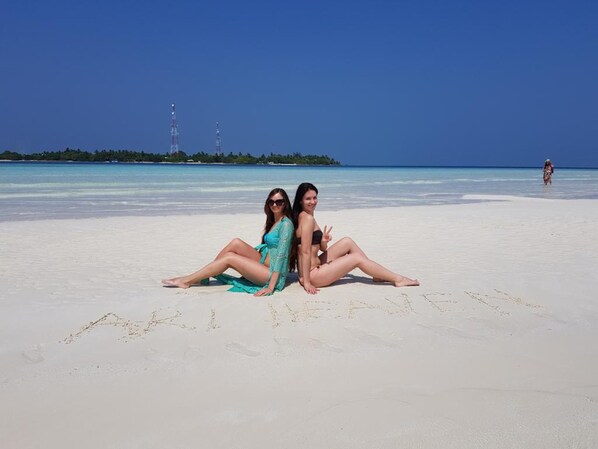 Deux personnes sont assises sur une plage de sable, avec un ciel bleu dégagé et une mer calme en arrière-plan.