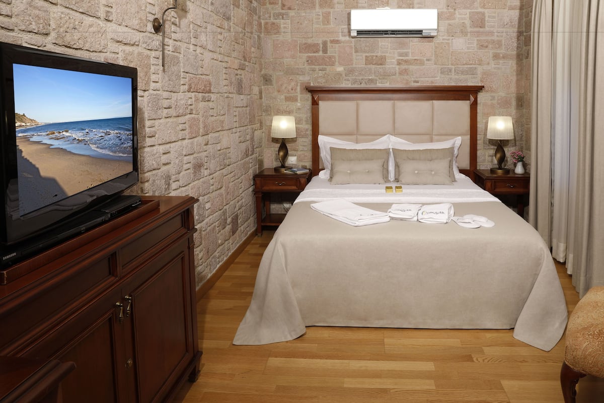 Ein Hotelzimmer mit Backsteinwand, einem Bett mit beiger Bettdecke, zwei Nachttischlampen, einem Flachbildfernseher auf einem Holzkasten und einem Holzboden.