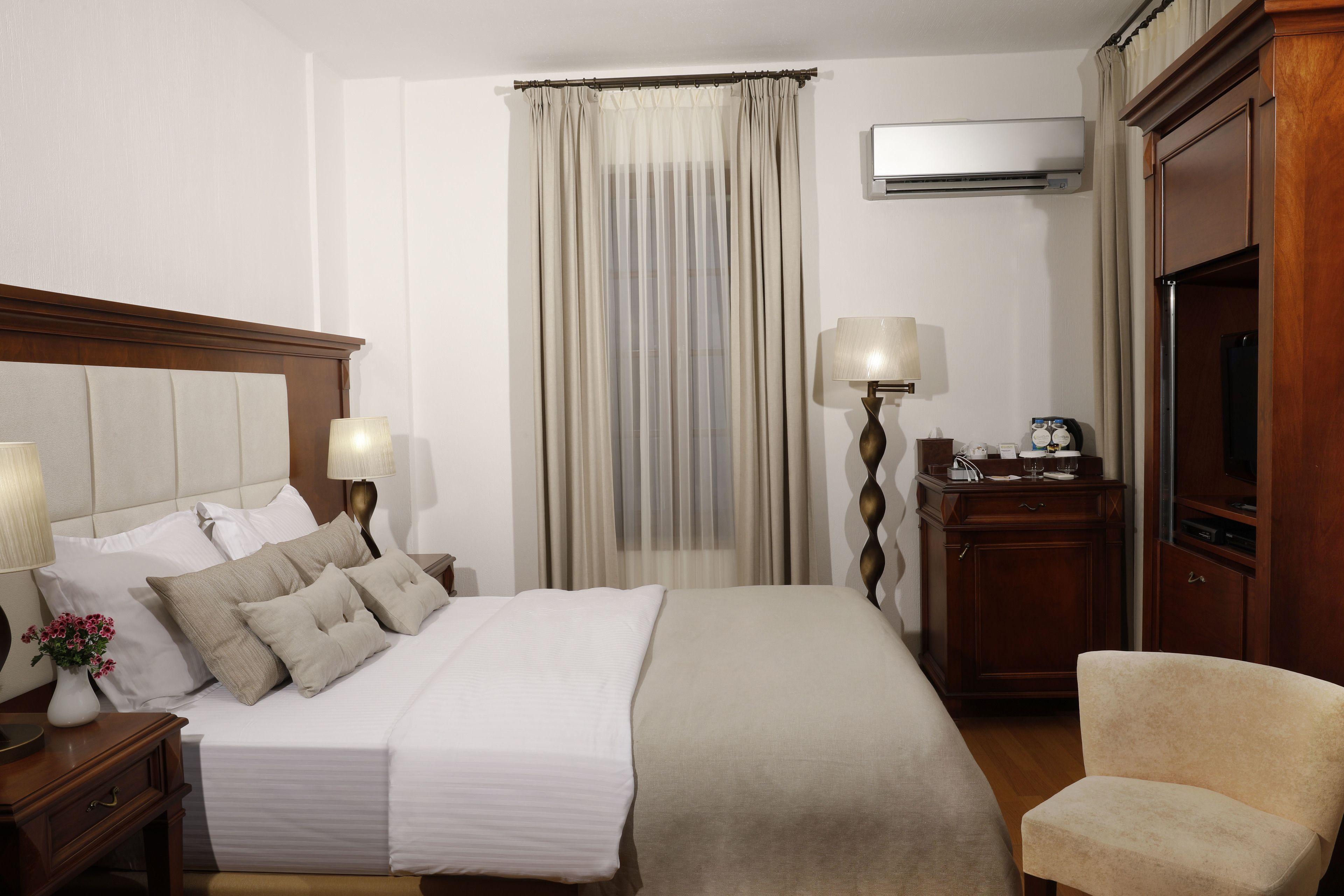 Habitación de hotel con una cama grande, lámparas de noche, una mesita de noche de madera, un escritorio con lámpara, un televisor y una silla.