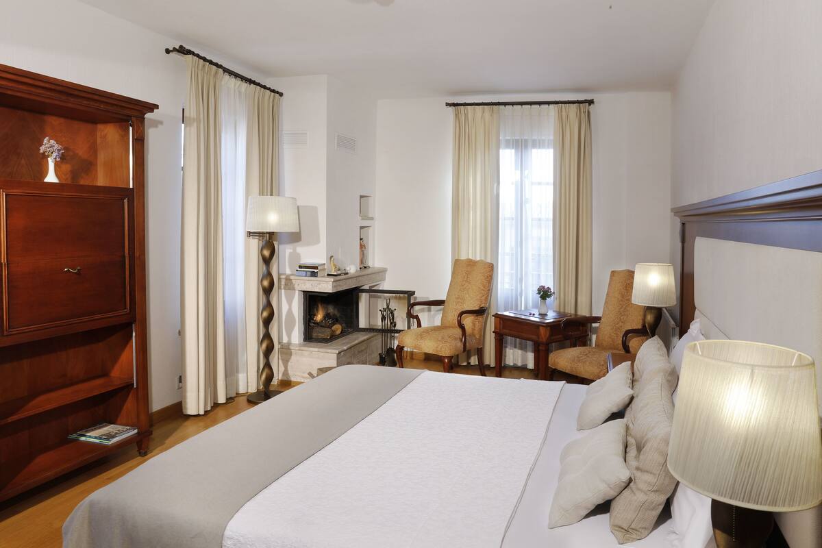 Ein Hotelzimmer mit einem großen Bett, einem hölzernen Kleiderschrank, einem Schreibtisch mit Lampe, zwei Sesseln und einem Kamin.