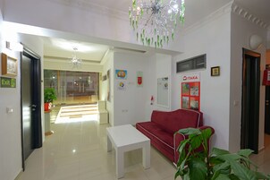 Una sala de estar moderna con una mesa de centro blanca, un sofá rojo y una araña. Hay una puerta de cristal que da a un balcón, y las paredes están decoradas con varios carteles e imágenes.