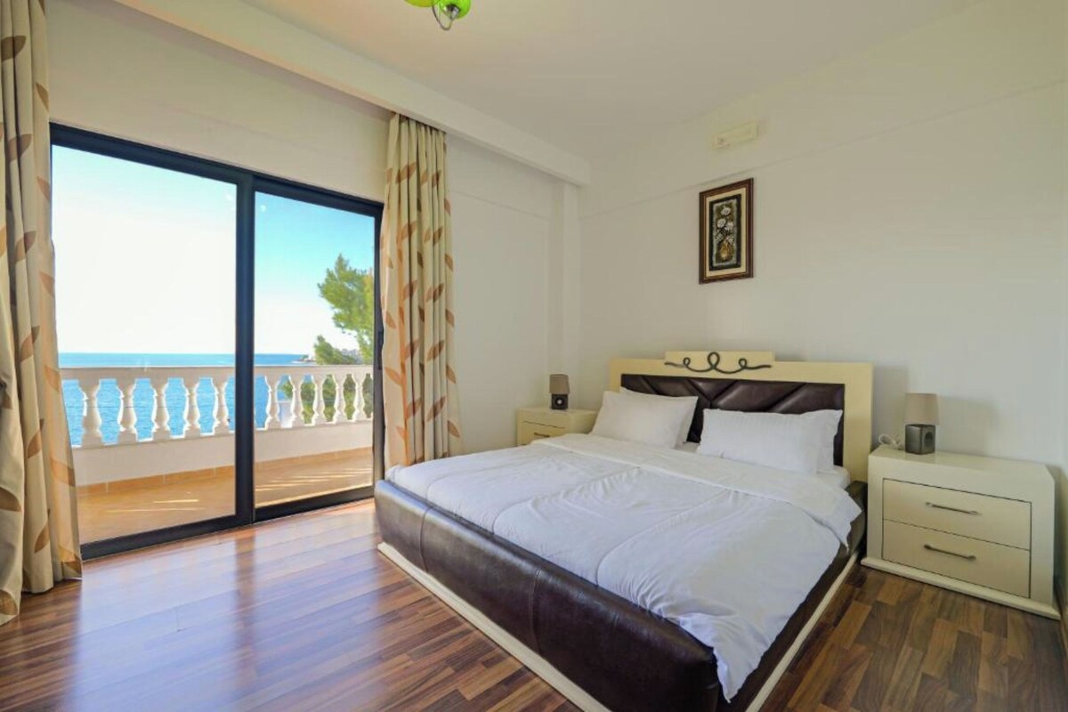 A bedroom with a large bed, a nightstand, a framed picture, and a view of the sea through a sliding door.