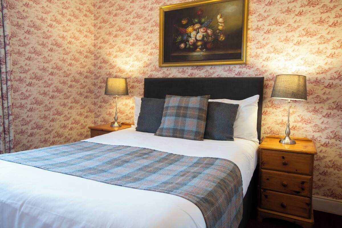 A neatly made bed with a plaid bedspread, two bedside lamps, a nightstand with drawers, and a floral-patterned wallpaper.