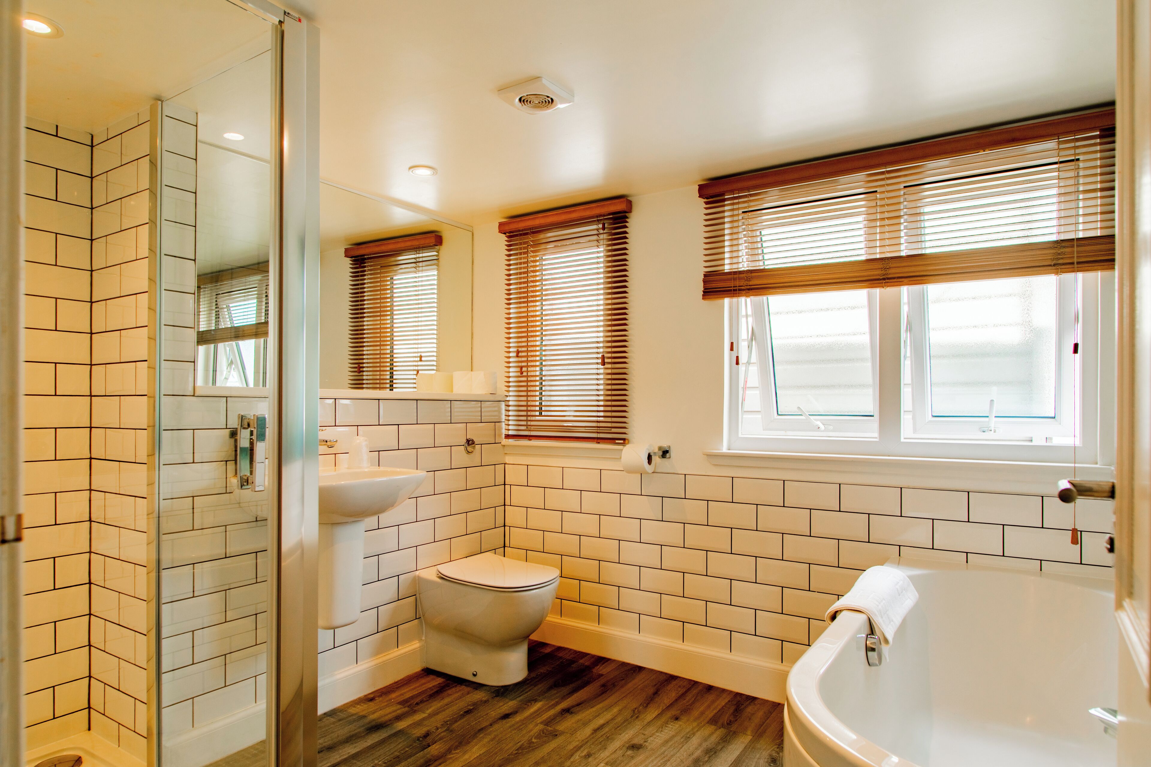 Un baño con inodoro, lavamanos y bañera, con persianas de madera y paredes de azulejos blancos.