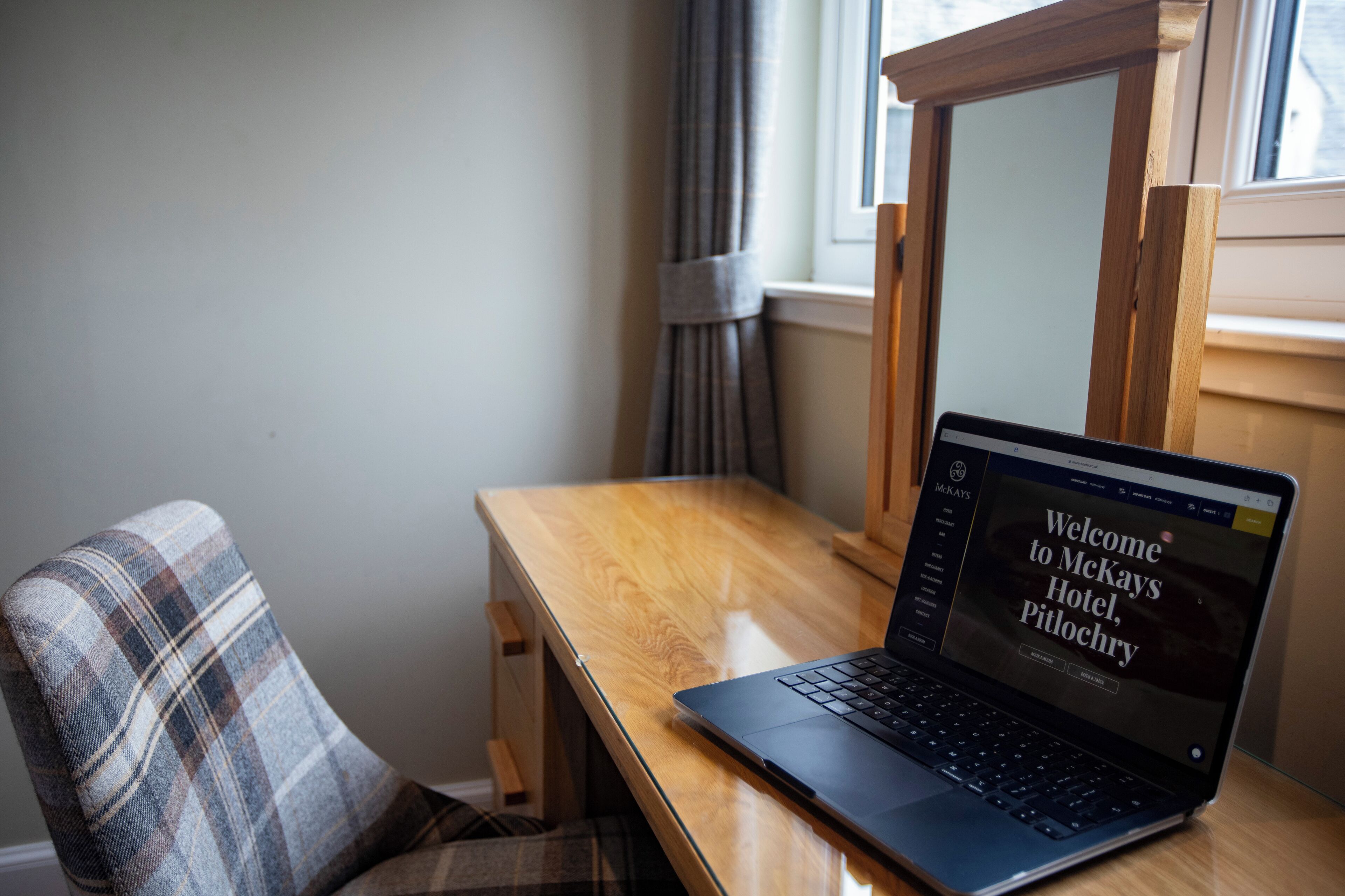 Un ordinateur portable est posé sur un bureau en bois, à côté d’une chaise à carreaux, d’une fenêtre avec des rideaux et d’un miroir.