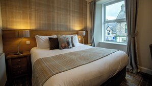 Una habitación de hotel con una cama grande, un cabecero con una lámpara, una mesita de noche con lámpara y una ventana con cortinas.