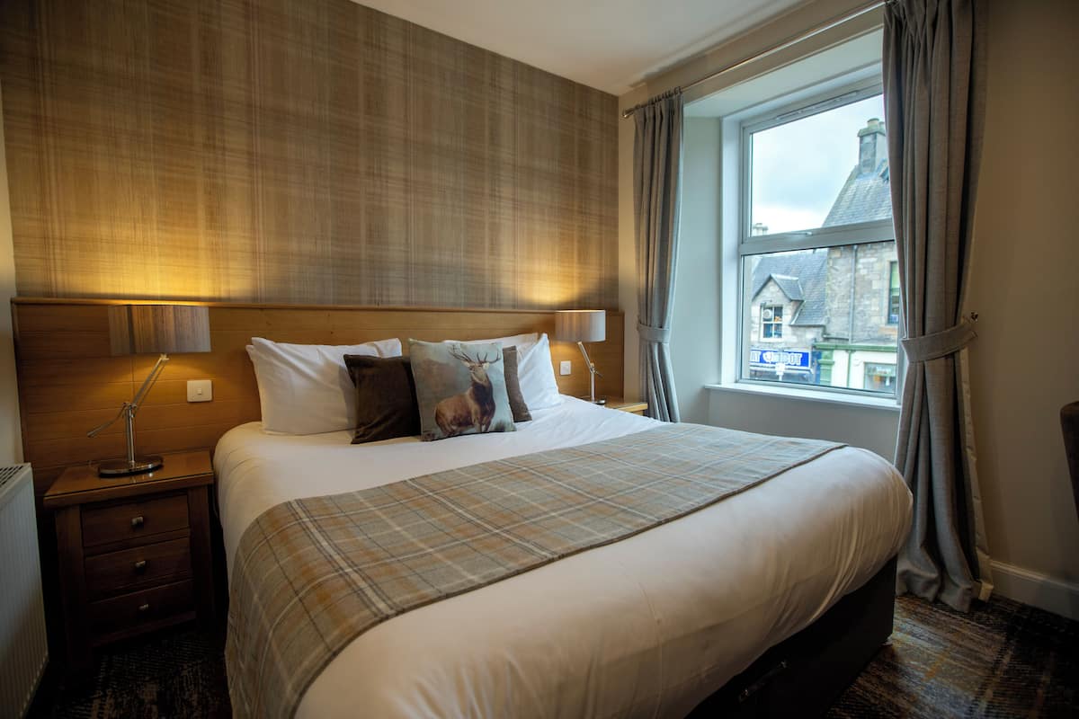 A hotel room with a large bed, a headboard with a light fixture, a bedside table with a lamp, and a window with curtains.