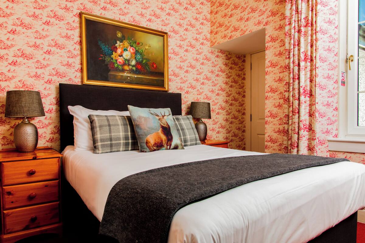 A neatly made bed with a floral wallpaper, a painting above it, and two lamps on either side.