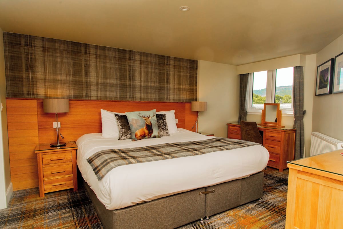 A hotel room with a large bed, wooden headboard, two bedside tables with lamps, a dresser, and a window with curtains.