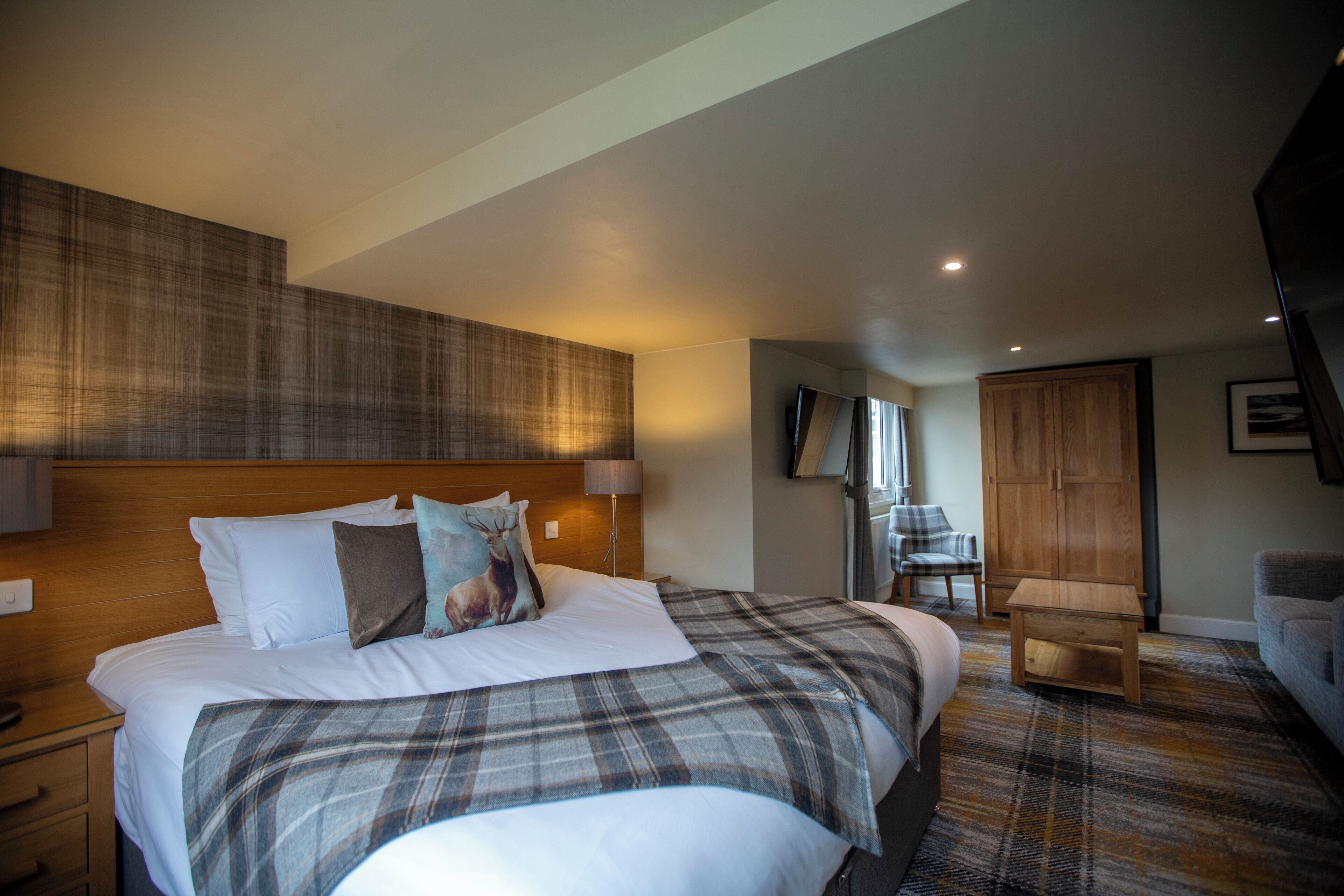 A modern hotel room with a large bed, a wooden headboard, a plaid bedspread, a small wooden nightstand, a lamp, a wooden wardrobe, and a television.