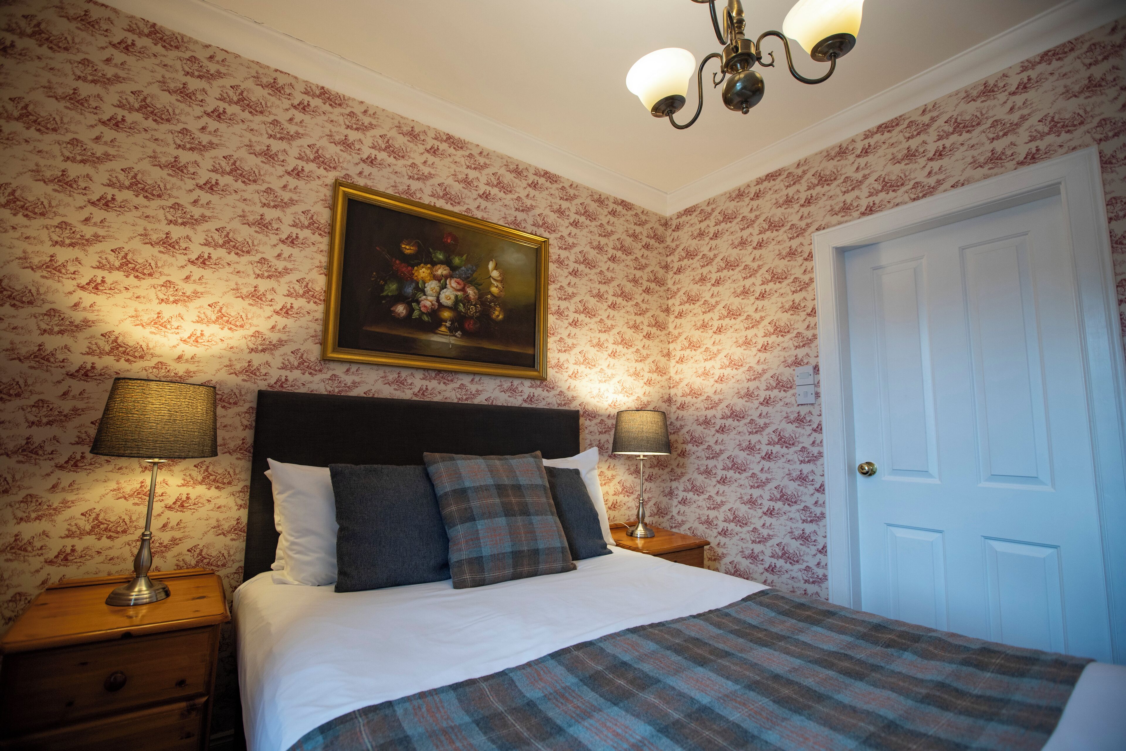 A neatly made bed with a plaid bedspread, two bedside lamps, a framed floral painting above the bed, and a white door to the right.