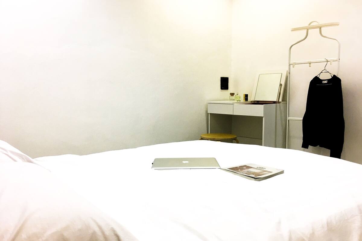 A neatly made bed with a laptop and a magazine, a small stool, a desk with a mirror, and a coat rack with a jacket.
