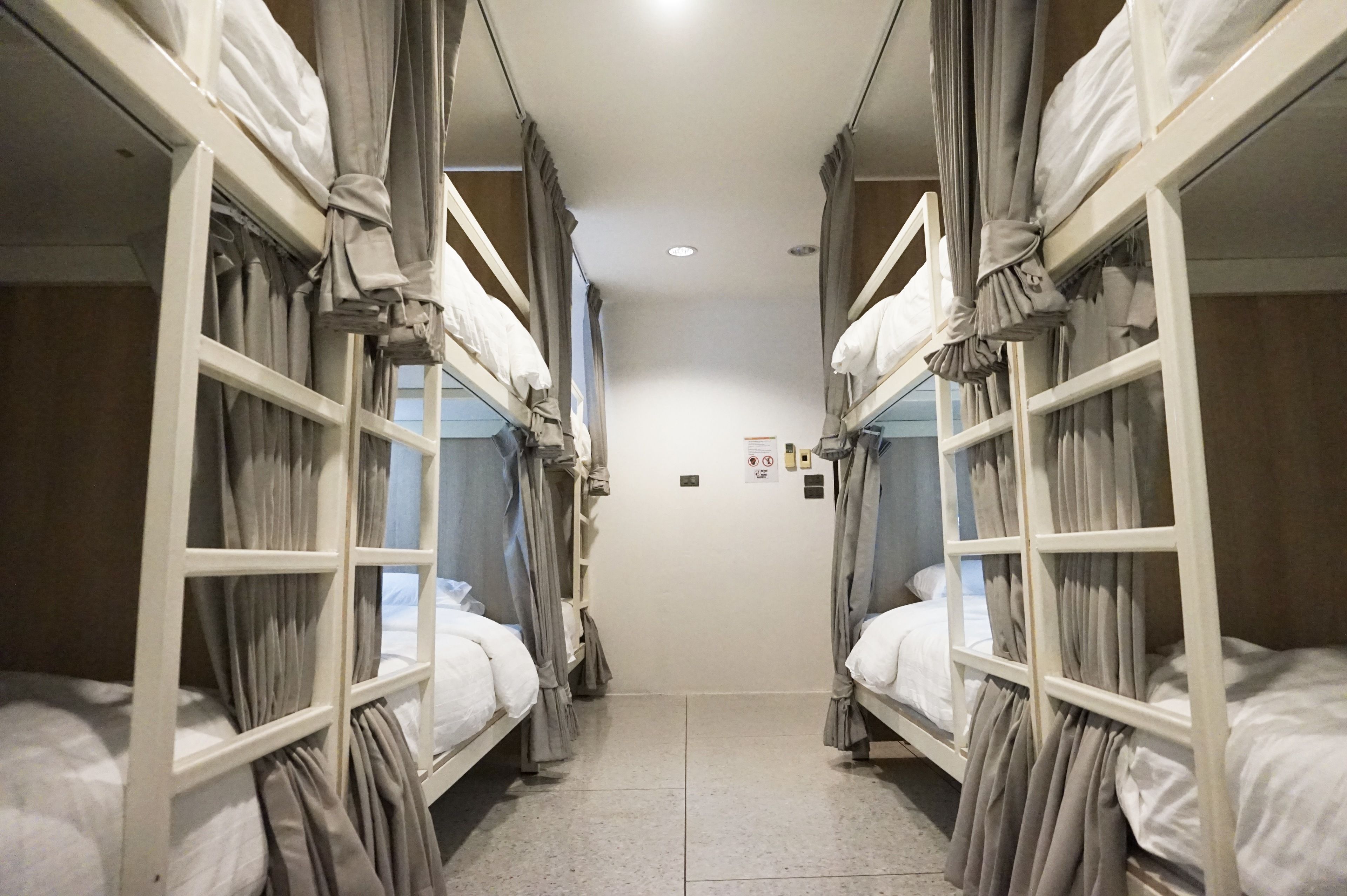 A narrow corridor with bunk beds on either side, a tiled floor, and a plain wall with a few signs.
