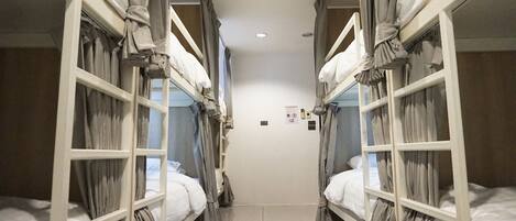 A narrow corridor with bunk beds on either side, a tiled floor, and a plain wall with a few signs.