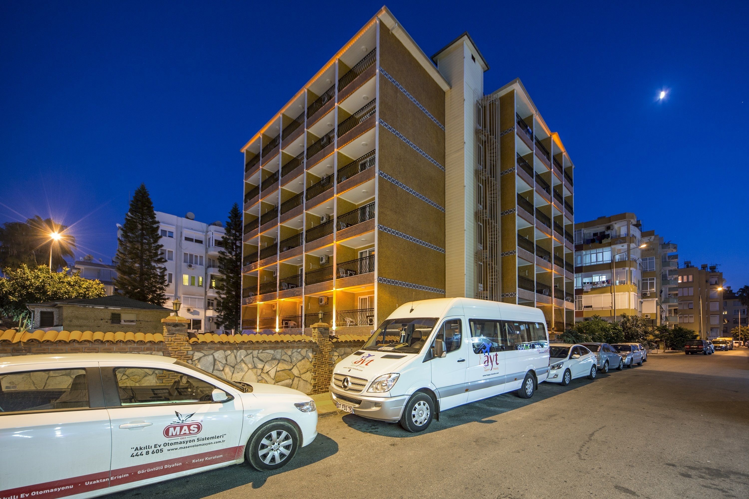 A multi-story building with a parking lot. There are several cars parked in front, including a white van with a logo. The building has balconies on each floor.
