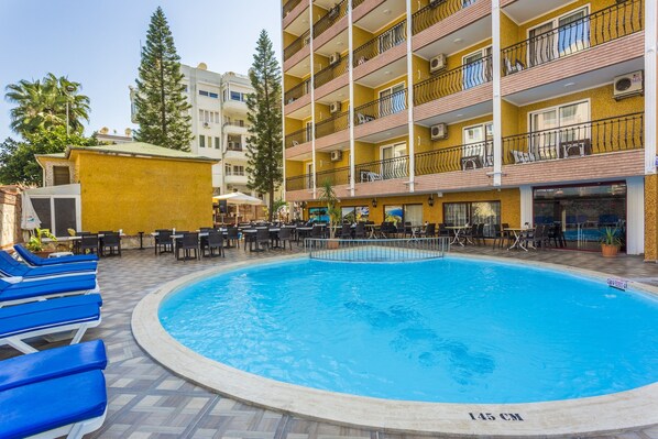 Zona de piscina de hotel con sillas reclinables azules, una piscina con indicador de profundidad y un edificio con balcones y unidades de aire acondicionado.