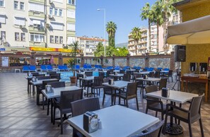 Un espacio de comedor al aire libre con mesas y sillas, piscina y edificios al fondo.