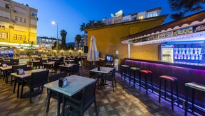 An outdoor dining area with tables and chairs, a bar with stools, and a view of the cityscape at dusk. - Wasa Hotel (Alanya)