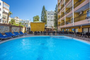 Una piscina con agua azul, rodeada de sillas de descanso y mesas, con edificios al fondo.