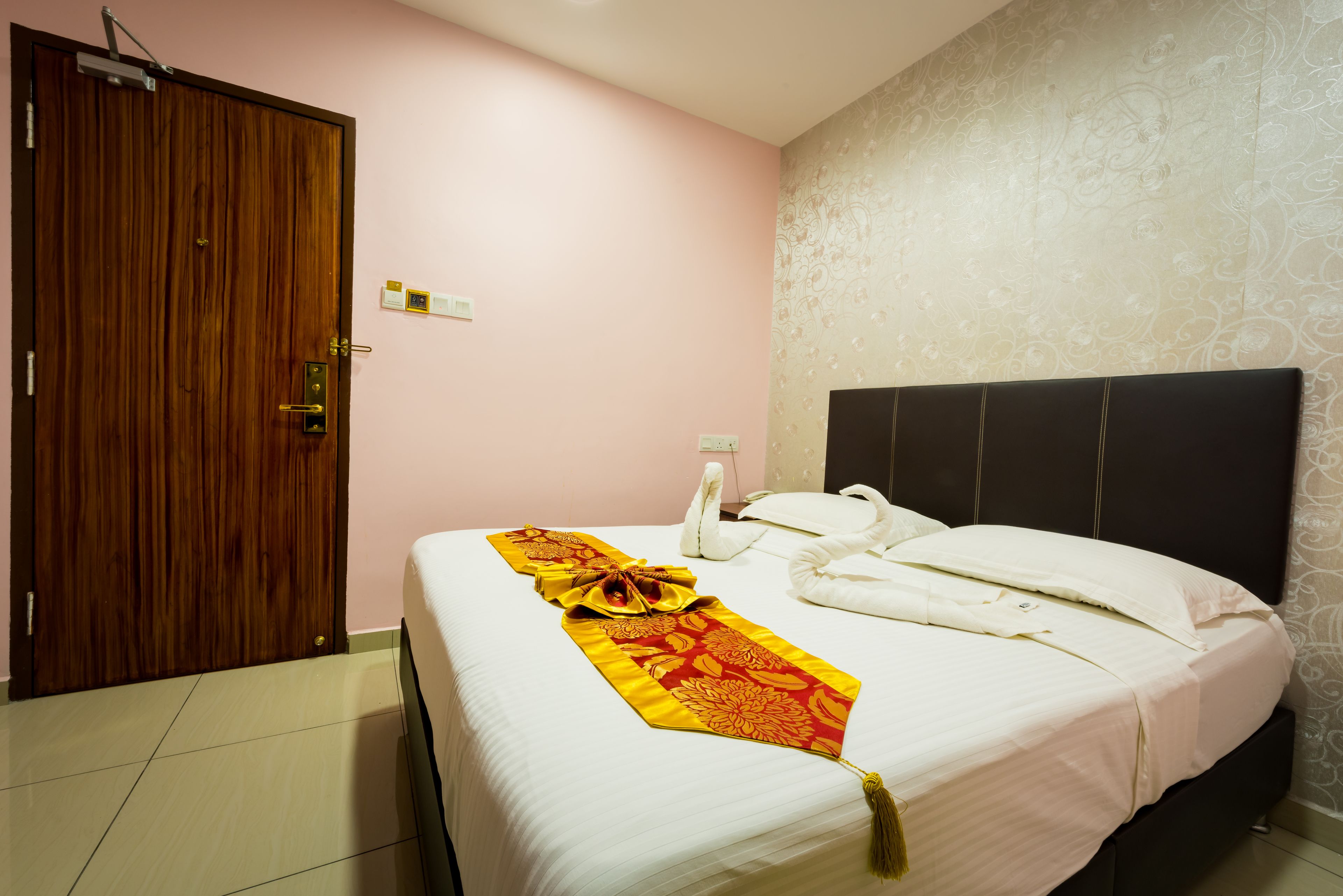 A hotel room with a wooden door, a bed with white linens, a decorative red and gold tie, and a black headboard.