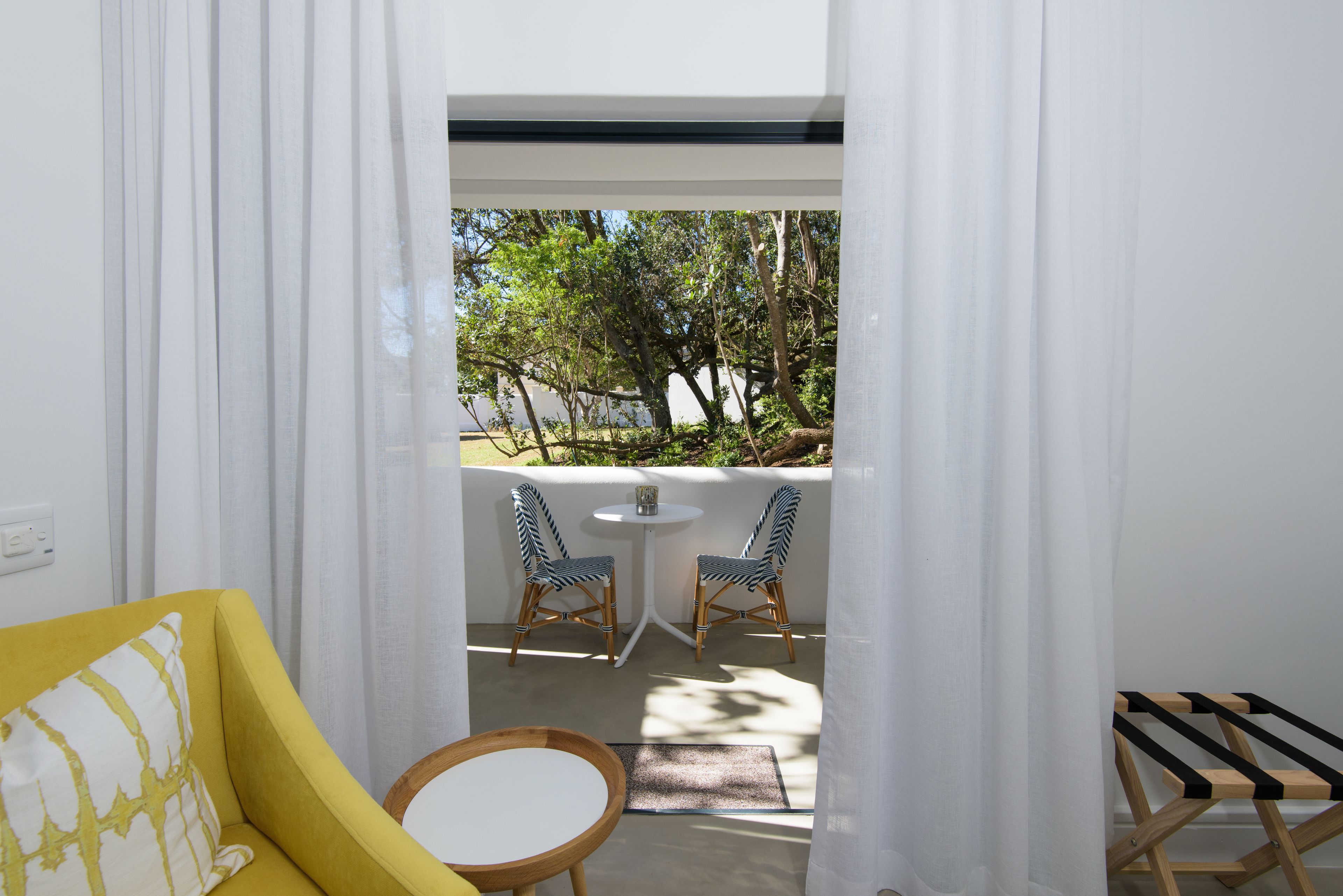 Habitación con sofá amarillo, una mesita redonda, dos sillas de mimbre y vistas a un balcón con mesa y sillas blancas.