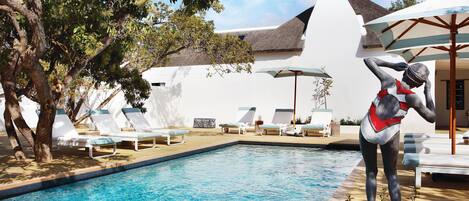 A swimming pool with lounge chairs and umbrellas, surrounded by trees and a building with a thatched roof.