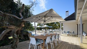 An outdoor patio with a wooden deck, white chairs, a wooden table, and large umbrellas providing shade. - The Old Rectory (Plettenberg Bay)