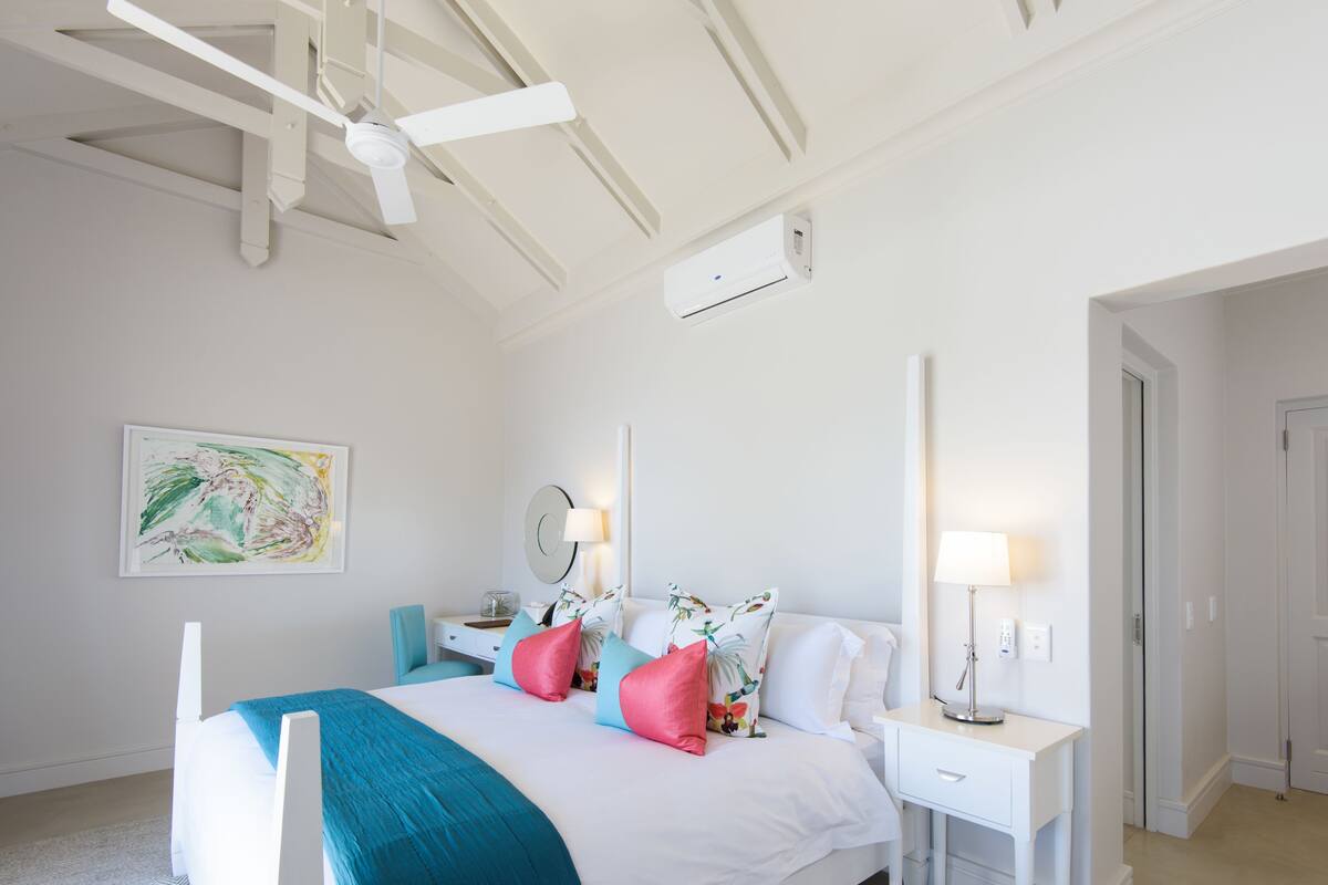 A bedroom with a bed, bedside tables, a ceiling fan, an air conditioning unit, a nightstand, a lamp, and a wall-mounted mirror.